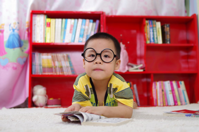 Child sitting at a table with a red bookcase behind him. Child is wearing a yellow and grey striped shirt with large round glasses on his face.