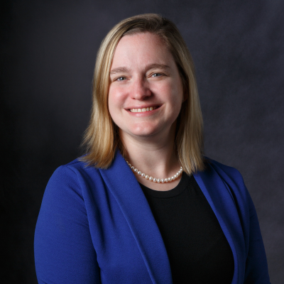 Professional photo of a person with a blue blazer, black shirt, and pearls on. She is smiling at the camera and her body is turned towards the left.