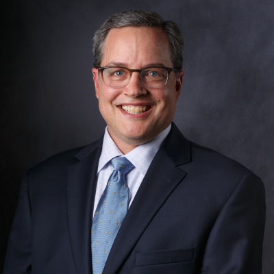 Professional photo of a person with glasses, a suit, dress shirt, and tie on. He is smiling at the camera and his body is turned towards his right.