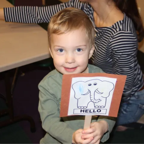 A little boy in a green shirt holding up a popsicle stick with a piece of paper with an elephant that says Hello