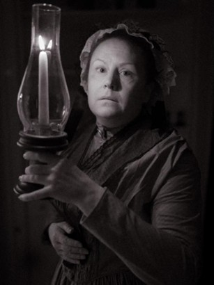 Photo of Laura F Keyes dressed as Lucy Pettengill standing in the dark holding a candle.